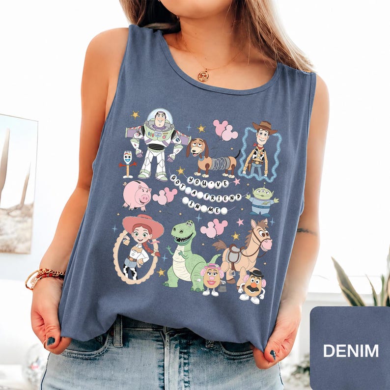 May include: A slate blue sleeveless tank top featuring a graphic print of various Toy Story characters including Buzz Lightyear, Woody, Jessie, Rex, Slinky Dog, and others, with the text "YOU'VE GOT A FRIEND IN ME" arranged in a circular pattern.