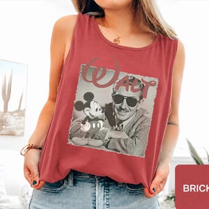 Peut inclure: Un d&eacute;bardeur sans manches rouge brique avec une impression graphique en noir et blanc de Walt Disney et Mickey Mouse. Le mot Walt est imprim&eacute; dans une grande police stylis&eacute;e au-dessus des personnages. Le d&eacute;bardeur a une encolure ronde.