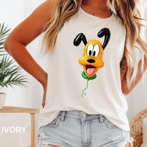 Op de afbeelding: Een ivoorkleurige mouwloze tanktop met een grafische print van een gele hond die is ontworpen als een glanzende ballon. Het personage heeft zwarte oren, een zwarte neus en een groene ballonknoop aan de onderkant met een dun groen touwtje.
