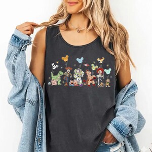 May include: Dark gray tank top featuring a colorful graphic of animated characters holding balloons. The characters include a green dinosaur, a cowboy, a space ranger, a horse, and a cowgirl. The balloons have various patterns and colors.