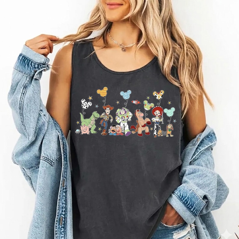 May include: Dark gray tank top featuring a colorful graphic of animated characters holding balloons. The characters include a green dinosaur, a cowboy, a space ranger, a horse, and a cowgirl. The balloons have various patterns and colors.