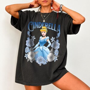 Puede incluir: Camiseta negra con un dise&ntilde;o de Cenicienta con un vestido azul, rodeada de flores azules y blancas. La palabra "CINDERELLA" est&aacute; impresa en azul encima de la imagen. La camiseta tiene mangas cortas y un corte holgado.