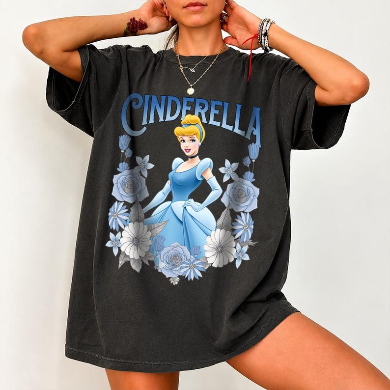 Puede incluir: Camiseta negra con un dise&ntilde;o de Cenicienta con un vestido azul, rodeada de flores azules y blancas. La palabra "CINDERELLA" est&aacute; impresa en azul encima de la imagen. La camiseta tiene mangas cortas y un corte holgado.