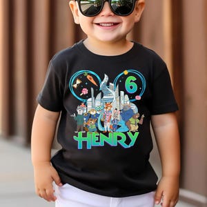 May include: Black t-shirt featuring a colorful graphic of cartoon characters, a cityscape, and the number 6. The name "HENRY" is displayed in a vibrant, multi-colored font. The shirt is designed for a child.