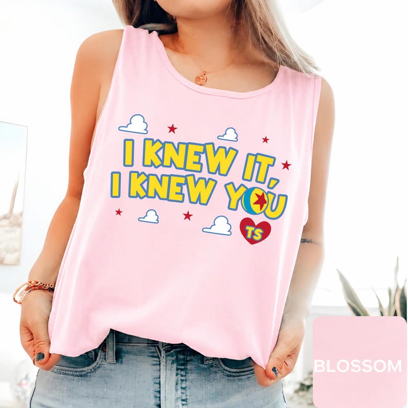 May include: A light pink sleeveless tank top featuring the yellow text "I KNEW IT, I KNEW YOU" with blue outlines. The design includes small white clouds, red stars, a red heart with the letters "TS" inside, and a colourful ball graphic.