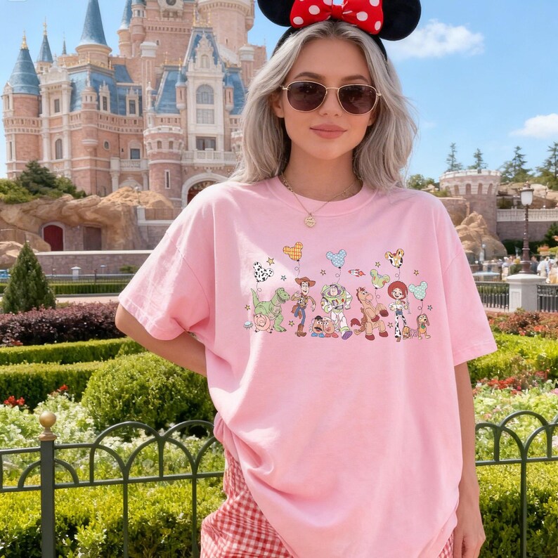 K&ouml;nnte beinhalten: Ein hellrosa T-Shirt mit einem Grafikdruck verschiedener animierter Spielzeugfiguren, die in einer Reihe stehen und jeweils einen bunten, mausf&ouml;rmigen Luftballon halten. Das T-Shirt hat kurze &Auml;rmel und einen Rundhalsausschnitt und wird zu einer rot karierten Hose getragen.
