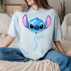 May include: A light blue t-shirt featuring a graphic of the character Stitch from Lilo and Stitch designed as a blue balloon with large pink-lined ears, big black eyes, and a blue string trailing downward with the word Stitch written in blue.