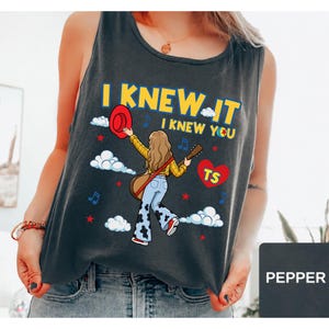 May include: A dark gray sleeveless tank top featuring a graphic illustration of a person with long hair wearing a yellow plaid shirt and cow-print bell-bottom jeans, holding a guitar and a red hat, with the text I KNEW IT I KNEW YOU and TS.