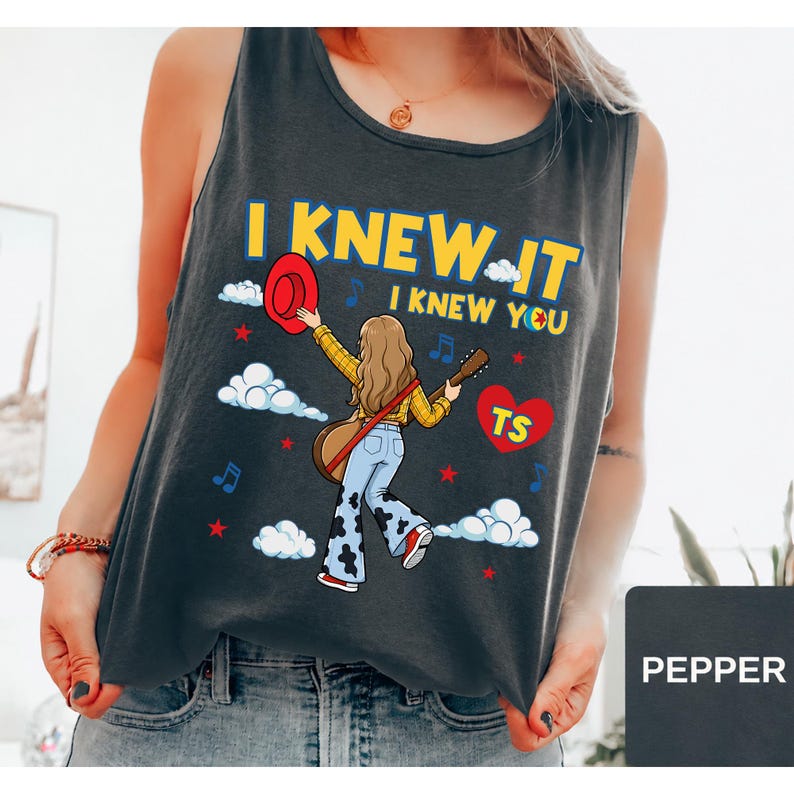 May include: A dark gray sleeveless tank top featuring a graphic illustration of a person with long hair wearing a yellow plaid shirt and cow-print bell-bottom jeans, holding a guitar and a red hat, with the text I KNEW IT I KNEW YOU and TS.