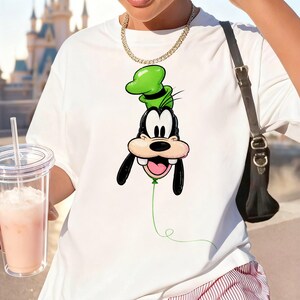 May include: A white short-sleeved t-shirt featuring a graphic print of the cartoon character Goofy designed as a green balloon with a thin green string extending downward. The shirt has a crew neckline and is worn with a gold chain necklace.