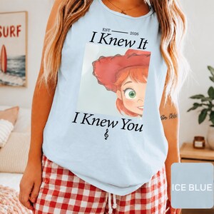 May include: A light blue sleeveless tank top featuring a graphic print of a cartoon character with red hair and a red hat. The text reads "EST 2026 I Knew It I Knew You" with a small musical note symbol below the text, centered on the chest.