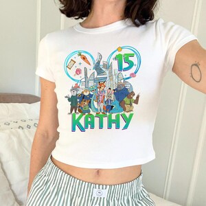 May include: White crop top with a colorful graphic featuring cartoon animals, the number 15, and the name "KATHY" in green and blue. The design includes a cityscape and various whimsical elements.