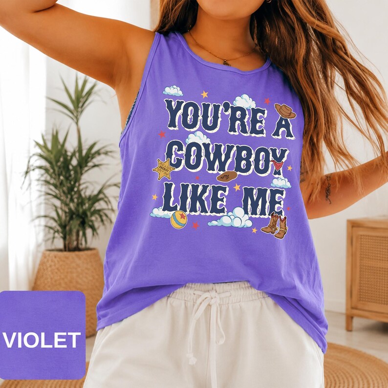 May include: A violet sleeveless tank top featuring the dark blue text 'YOU'RE A COWBOY LIKE ME' surrounded by graphic illustrations of a sheriff badge, a cowboy hat, a ball, and brown cowboy boots, accented with small white clouds and stars.