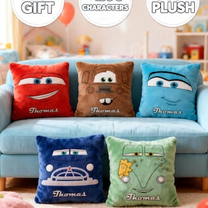 May include: Five plush character pillows in red, brown, blue, dark blue, and green, each with a different cartoon car face. The pillows are on a light blue couch. Each pillow has the name "Thomas" embroidered on it.