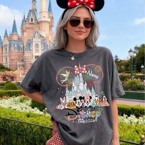 May include: Dark gray t-shirt featuring a Disney theme park design. The graphic includes Disney characters, a castle, fireworks, and the text "Disney Trip 2020". The design is framed by a pink bow and the word "Name".