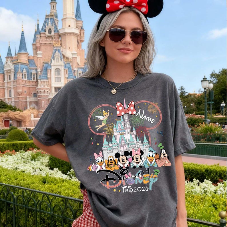 May include: Dark gray t-shirt featuring a Disney theme park design. The graphic includes Disney characters, a castle, fireworks, and the text "Disney Trip 2020". The design is framed by a pink bow and the word "Name".