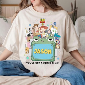 May include: Cream-colored t-shirt featuring a colorful graphic of characters from Toy Story 5. A green frog holds a blue sign with the name "JASON". The text "YOU'VE GOT A FRIEND IN ME" is below the frog. Stars and other characters surround the main image.