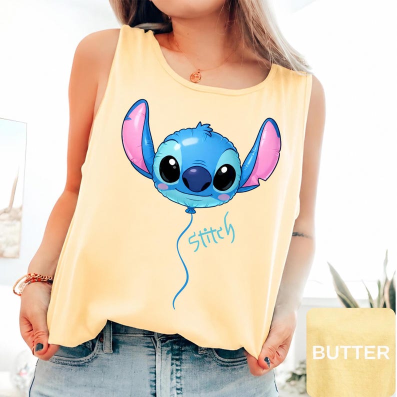 May include: A pale yellow sleeveless tank top featuring a graphic of a blue character head shaped like a balloon with large black eyes and pink inner ears. A thin blue string extends downward from the balloon with the word Stitch written in blue.