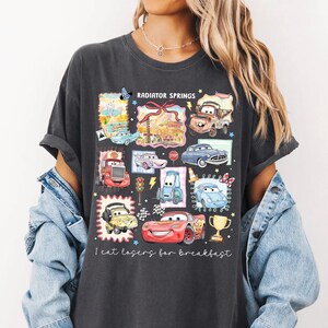 May include: Dark gray t-shirt featuring a colorful graphic of cartoon cars and the text "Radiator Springs" and "I eat losers for breakfast". The design includes various car characters and a trophy.