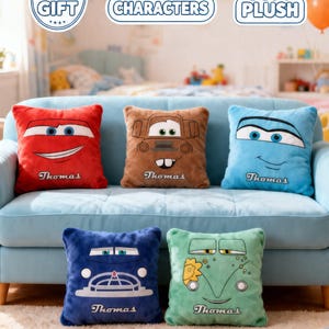 May include: Five plush square pillows with car-themed designs in red, brown, blue, dark blue, and green. Each pillow features a car face and the name "Thomas" below. The pillows are on a light blue couch.