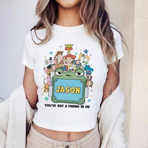 May include: White crop top featuring a colorful graphic of Toy Story characters. The design includes a frog holding a sign that says "JASON" and the phrase "YOU'VE GOT A FRIEND IN ME". The top has short sleeves and a crew neck.