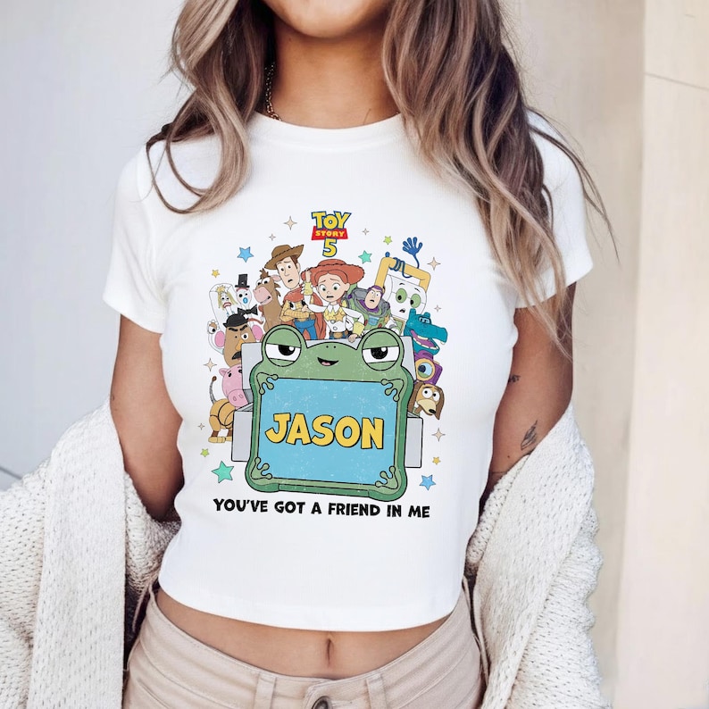 May include: White crop top featuring a colorful graphic of Toy Story characters. The design includes a frog holding a sign that says "JASON" and the phrase "YOU'VE GOT A FRIEND IN ME". The top has short sleeves and a crew neck.
