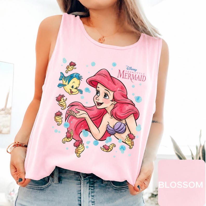 Peut inclure: Un d&eacute;bardeur rose clair sans manches avec une impression graphique d'Ariel de La Petite Sir&egrave;ne aux cheveux rouges, un haut coquillage violet et le poisson Polochon. Le motif inclut des petits hippocampes jaunes et des bulles bleues avec le texte Disney The Little Mermaid.