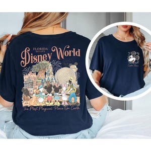 May include: Navy blue t-shirt featuring a Disney World design. The back of the shirt displays the words "Disney World Florida" with a graphic of Disney characters and landmarks. The front has a small Mickey Mouse graphic. The text "The Most Magical Place On Earth" is also visible.