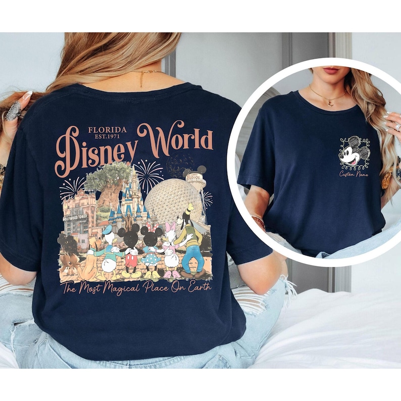 May include: Navy blue t-shirt featuring a Disney World design. The back of the shirt displays the words "Disney World Florida" with a graphic of Disney characters and landmarks. The front has a small Mickey Mouse graphic. The text "The Most Magical Place On Earth" is also visible.
