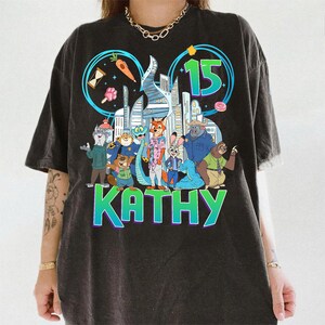 May include: Dark gray t-shirt featuring a colorful graphic of characters from the movie Zootopia, with the number 15 and the name KATHY. The design includes a cityscape and various food items. The shirt has a vintage, worn look.