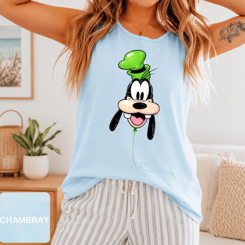 Op de afbeelding: Een lichtblauw mouwloos hemdje met een grafische print van het personage Goofy ontworpen als een groene ballon. Het personage heeft een zwarte neus, lange zwarte oren en een groene hoed. Het woord CHAMBRAY staat in een klein vakje onderaan.