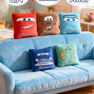 May include: A collection of plush pillows in various colors, each featuring a cartoon car face. The pillows are red, brown, blue, and green, with the name "Thomas" embroidered on each. They are arranged on a light blue sofa.