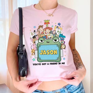 May include: A light pink t-shirt featuring a colorful graphic from the movie "Toy Story 5". The design includes various characters, the name "JASON", and the phrase "YOU'VE GOT A FRIEND IN ME".