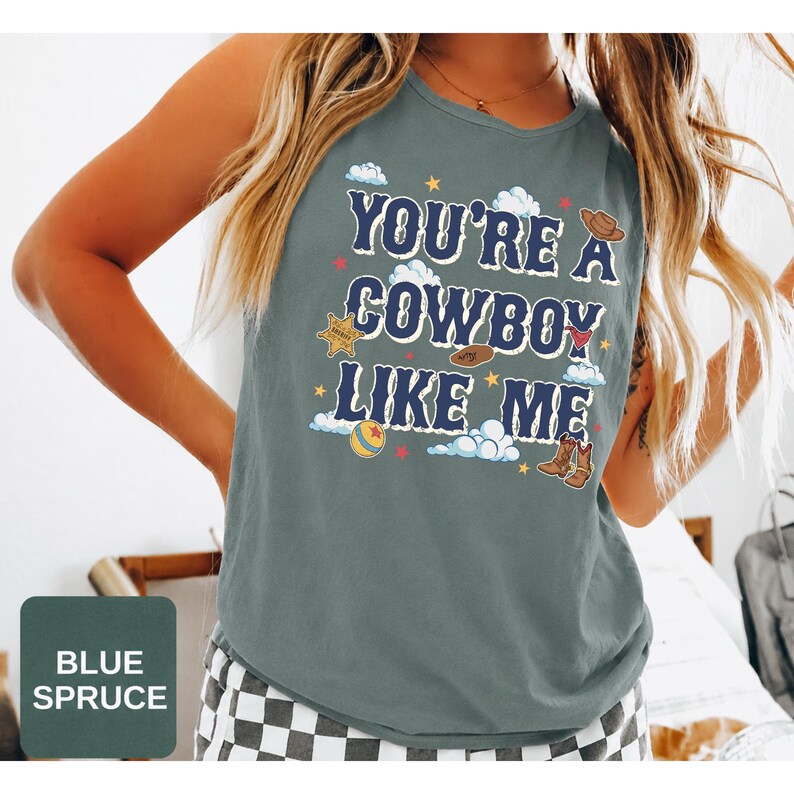 May include: A blue spruce sleeveless tank top featuring the text "YOU'RE A COWBOY LIKE ME" in dark blue letters. The graphic includes illustrations of a sheriff badge, a cowboy hat, a red bandana, a ball, and brown cowboy boots with clouds and stars.