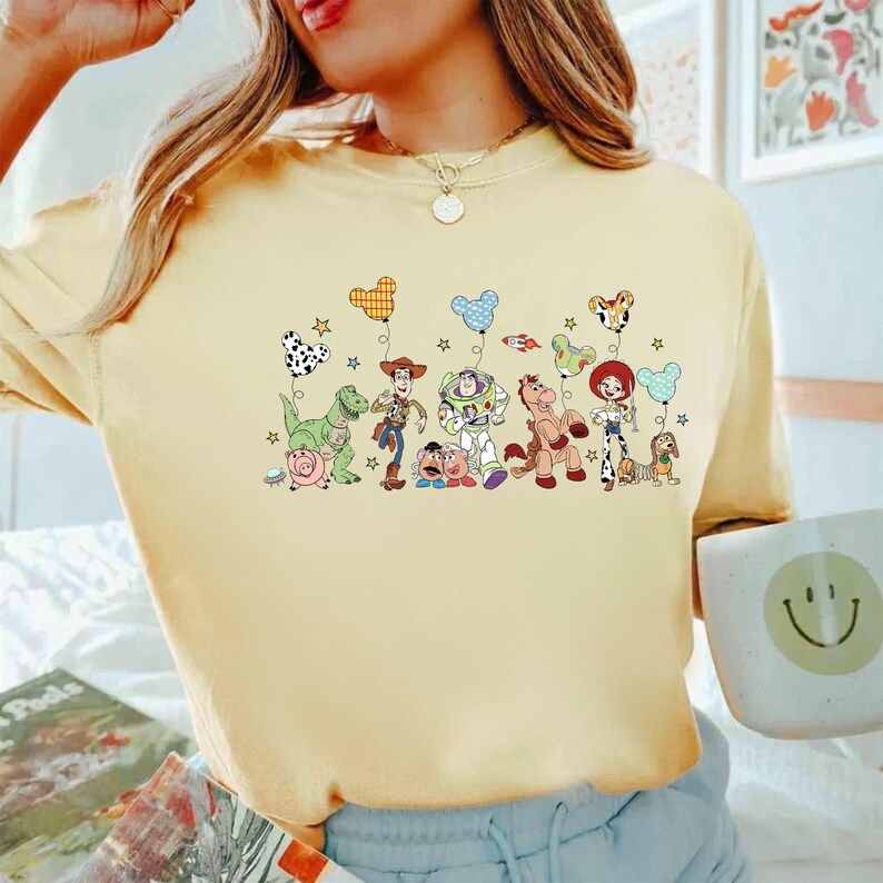 K&ouml;nnte beinhalten: Ein blassgelbes T-Shirt mit Rundhalsausschnitt und einem Grafikdruck verschiedener animierter Figuren aus der Toy Story-Reihe. Die Figuren halten bunte Luftballons in Form von Mickey-Maus-Ohren, begleitet von kleinen Sternen und einer Rakete.