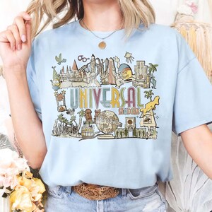 May include: Pale blue t-shirt with a vibrant graphic of Universal Studios attractions and the words "UNIVERSAL STUDIOS". The design includes cartoon characters and landmarks, ideal for theme park fans.