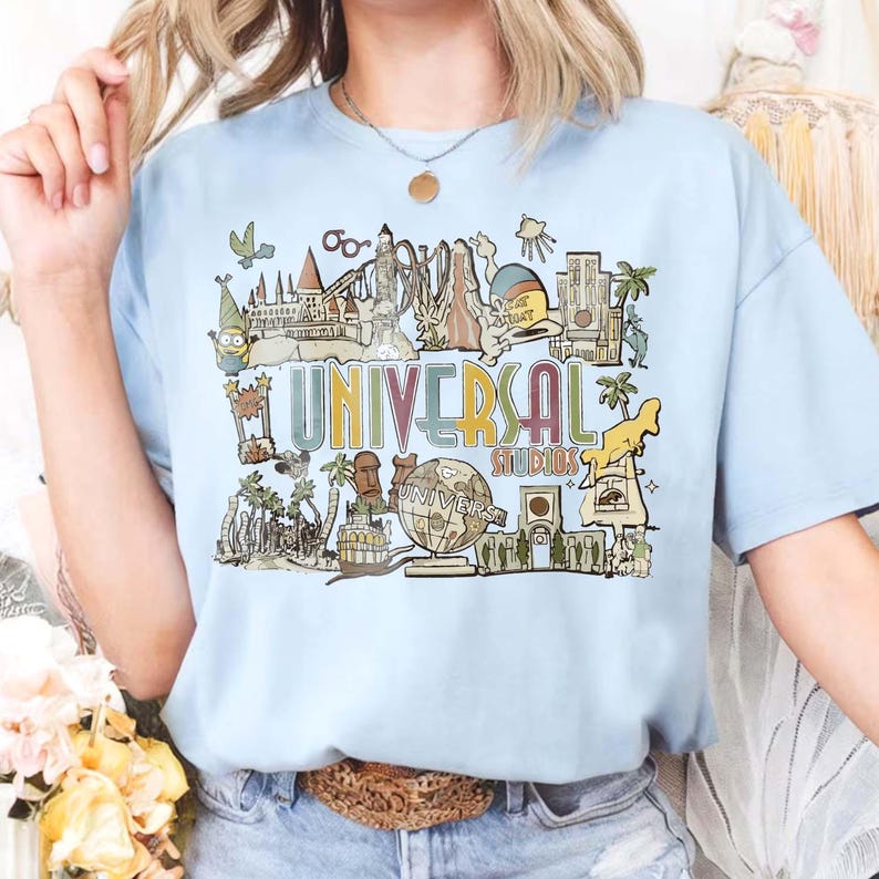 May include: Pale blue t-shirt with a vibrant graphic of Universal Studios attractions and the words "UNIVERSAL STUDIOS". The design includes cartoon characters and landmarks, ideal for theme park fans.