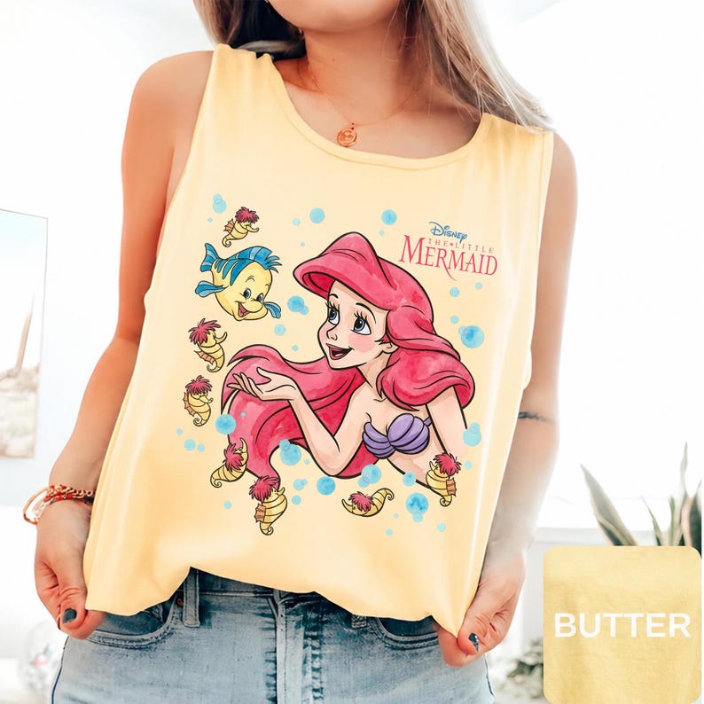 Peut inclure: Un d&eacute;bardeur jaune p&acirc;le sans manches avec une impression graphique d'Ariel de La Petite Sir&egrave;ne aux cheveux rouges, un haut coquillage violet et Polochon. Le motif inclut des hippocampes et des bulles bleues avec le texte Disney The Little Mermaid.