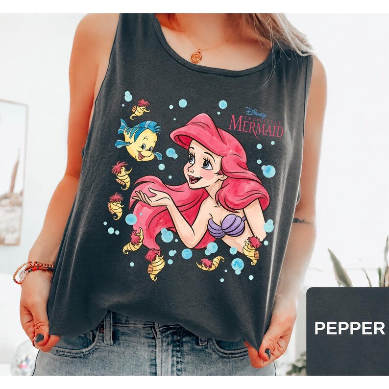Peut inclure: Un d&eacute;bardeur gris anthracite sans manches avec une impression graphique d'Ariel de La Petite Sir&egrave;ne aux longs cheveux rouges, un haut en coquillages violets et le poisson Polochon. Le motif inclut des bulles bleues et le texte Disney The Little Mermaid.