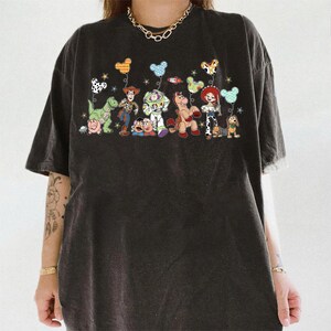 K&ouml;nnte beinhalten: Ein anthrazitfarbenes T-Shirt mit einem Grafikdruck verschiedener animierter Spielzeugfiguren, darunter ein Cowboy, ein Weltraum-Ranger, ein Cowgirl, ein Dinosaurier, ein Schwein, eine Kartoffelfigur, ein Pferd und ein Dackel mit Luftballons.