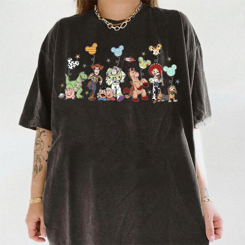 K&ouml;nnte beinhalten: Ein anthrazitfarbenes T-Shirt mit einem Grafikdruck verschiedener animierter Spielzeugfiguren, darunter ein Cowboy, ein Weltraum-Ranger, ein Cowgirl, ein Dinosaurier, ein Schwein, eine Kartoffelfigur, ein Pferd und ein Dackel mit Luftballons.