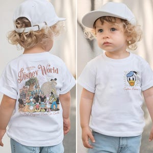 May include: White t-shirt with a Disney World graphic on the back and a Donald Duck design on the front. The shirt has rolled-up sleeves and is worn with a white baseball cap. The text "Custom Name" is below the Donald Duck graphic.