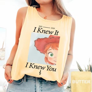 May include: A pale yellow sleeveless tank top featuring a graphic print of a cartoon character with red hair and a red hat. The text reads EST 2026 I Knew It I Knew You with a treble clef symbol below. The design is centered on the front of the shirt.