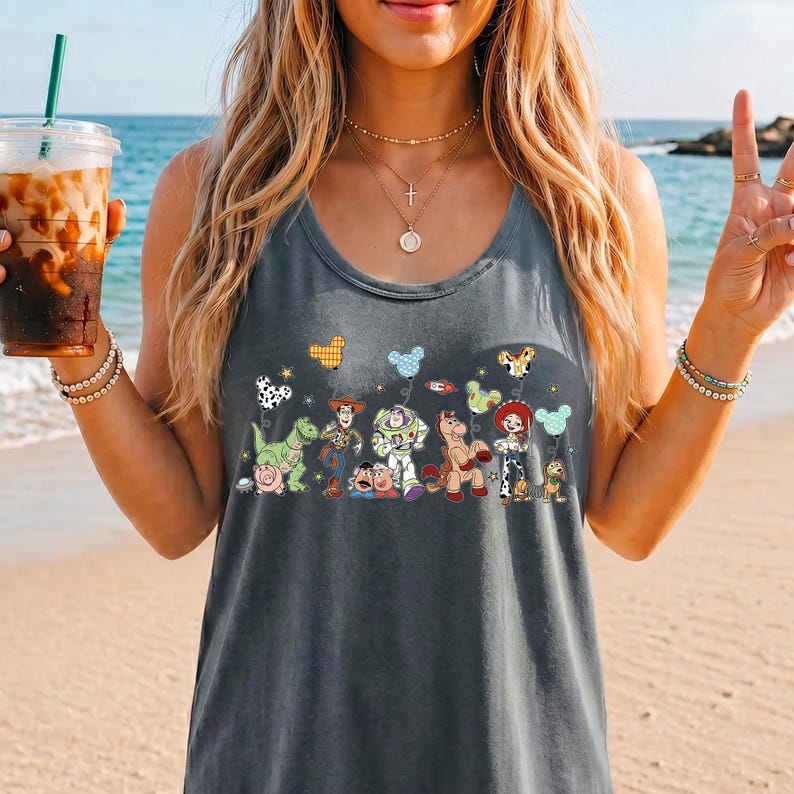 May include: Gray tank top featuring a colorful graphic of characters from the animated movie Toy Story. The design includes images of Woody, Buzz Lightyear, and other characters. The tank top is sleeveless and made of a soft material.