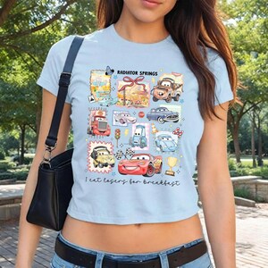 May include: Light blue t-shirt featuring a colorful graphic of cartoon cars and the text "Radiator Springs" and "I eat losers for breakfast". The design includes various car characters and racing elements.