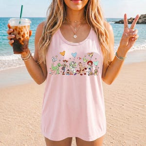 May include: A light pink tank top featuring a colorful graphic of cartoon characters. The design includes a green dinosaur, a cowboy, a space ranger, a horse, and other characters. The tank top is sleeveless and has a round neckline.