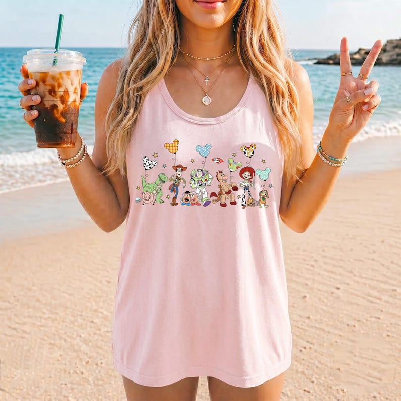 May include: A light pink tank top featuring a colorful graphic of cartoon characters. The design includes a green dinosaur, a cowboy, a space ranger, a horse, and other characters. The tank top is sleeveless and has a round neckline.