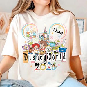 Peut inclure: T-shirt cr&egrave;me avec un graphisme color&eacute; de Disney World. Le motif comprend le ch&acirc;teau de Disney World, divers personnages de dessins anim&eacute;s et le texte "Disneyworld 2020". Un "Nom" personnalisable est &eacute;galement inclus.