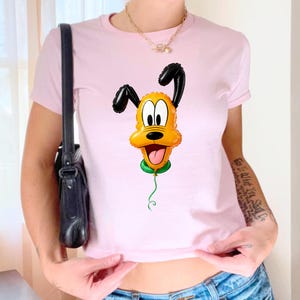 May include: A light pink short-sleeved t-shirt featuring a graphic print of a yellow dog character designed to look like a shiny, inflated foil balloon. The character has black ears, a green collar, and a thin green string hanging downward.