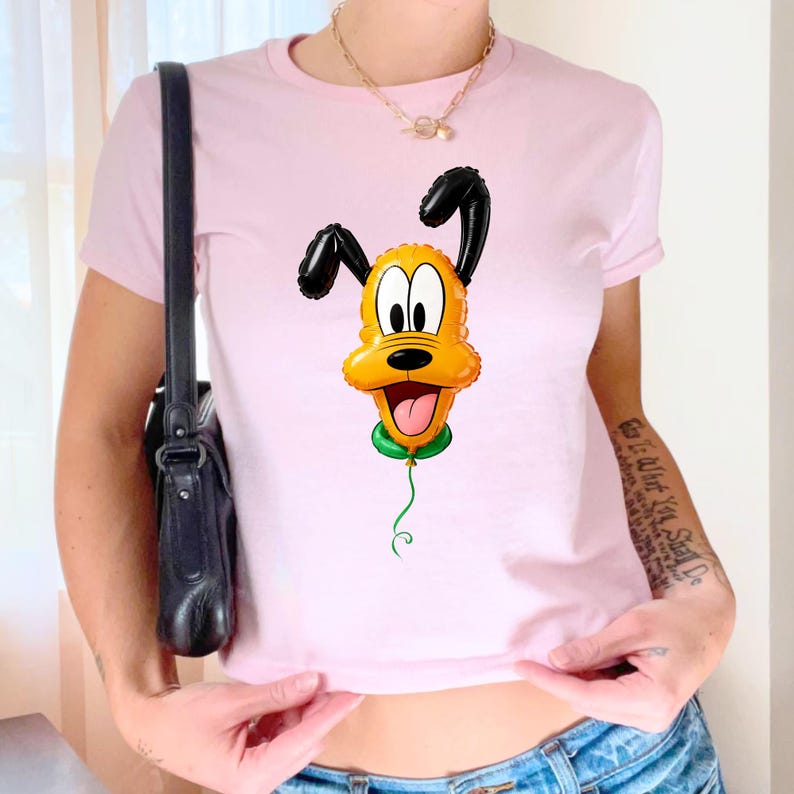 May include: A light pink short-sleeved t-shirt featuring a graphic print of a yellow dog character designed to look like a shiny, inflated foil balloon. The character has black ears, a green collar, and a thin green string hanging downward.